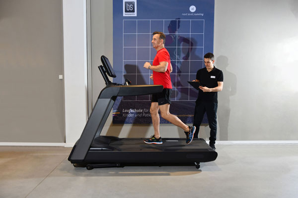 Ein Mann läuft auf einem Laufband, ein Trainer analysiert mit Tablet die Bewegungsausführung