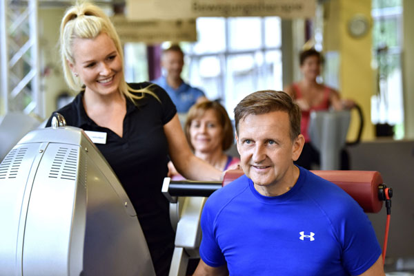 Ein Mann trainiert an einem Trainingsgerät, eine Physiotherapeutin in schwarzer Kleidung begleitet ihn, weitere Personen im Hintergrund