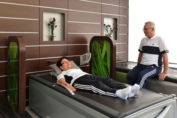 Person liegt entspannt auf einem therapeutischen Massagegerät, betreut von einem Begleiter